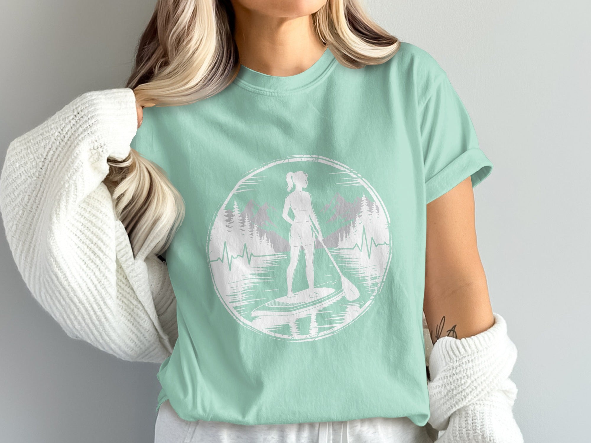 Woman wearing a mint t-shirt with a paddleboarding graphic and mountain backdrop.