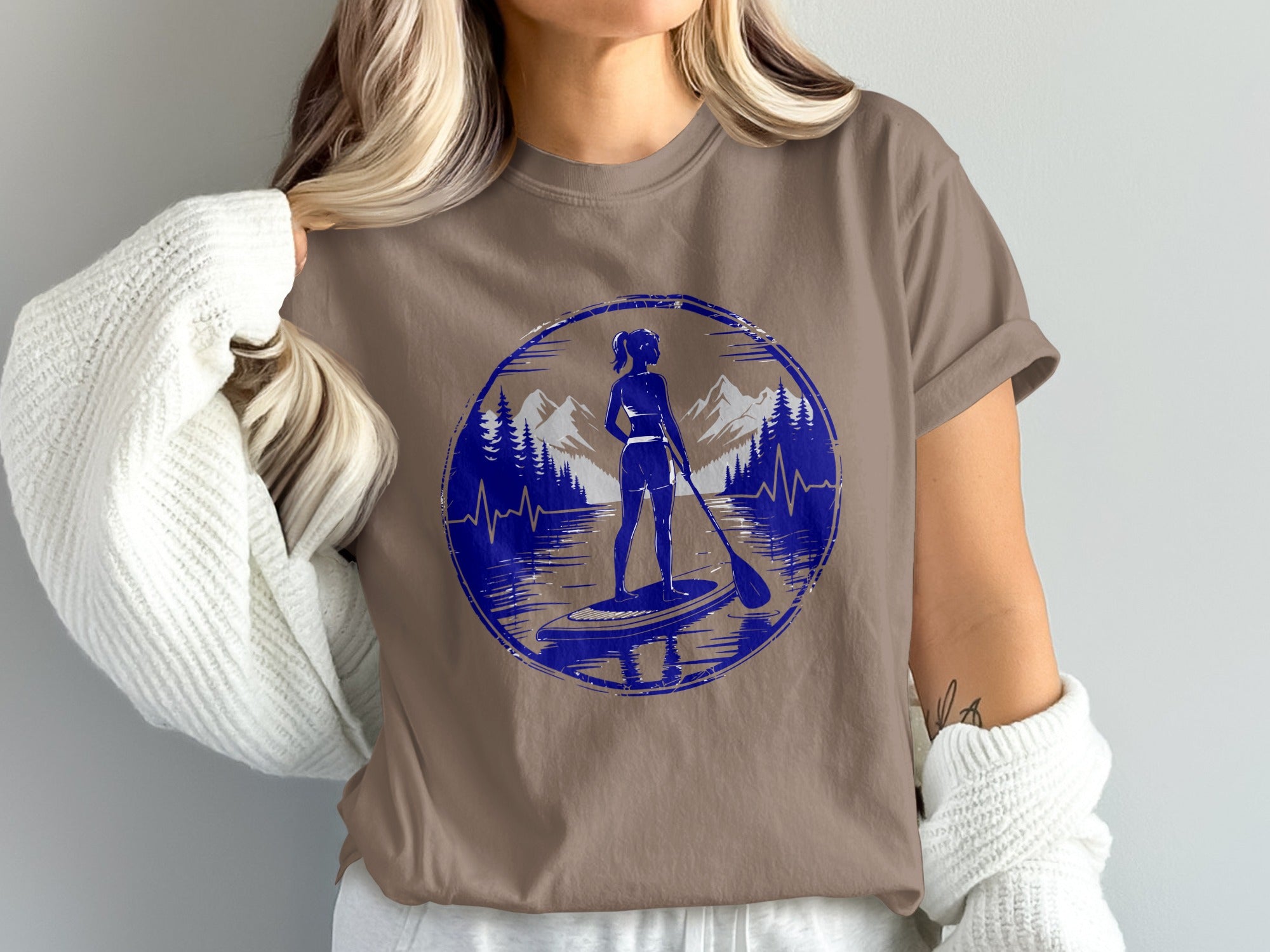 Woman on paddleboard with mountains and trees graphic on brown t-shirt.