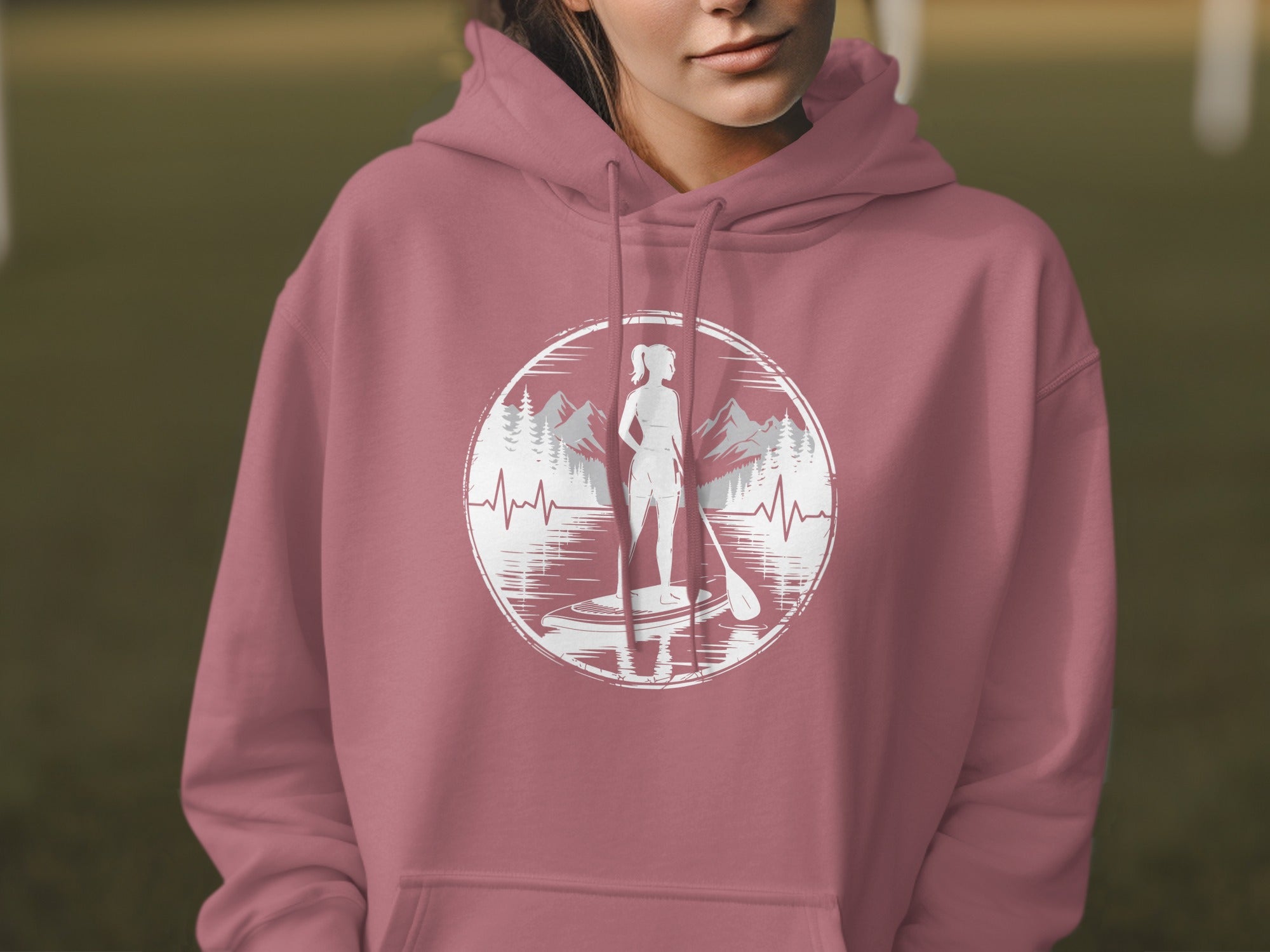 Woman in pink hoodie wearing graphic of paddleboarder on water with mountains and heartbeat line.