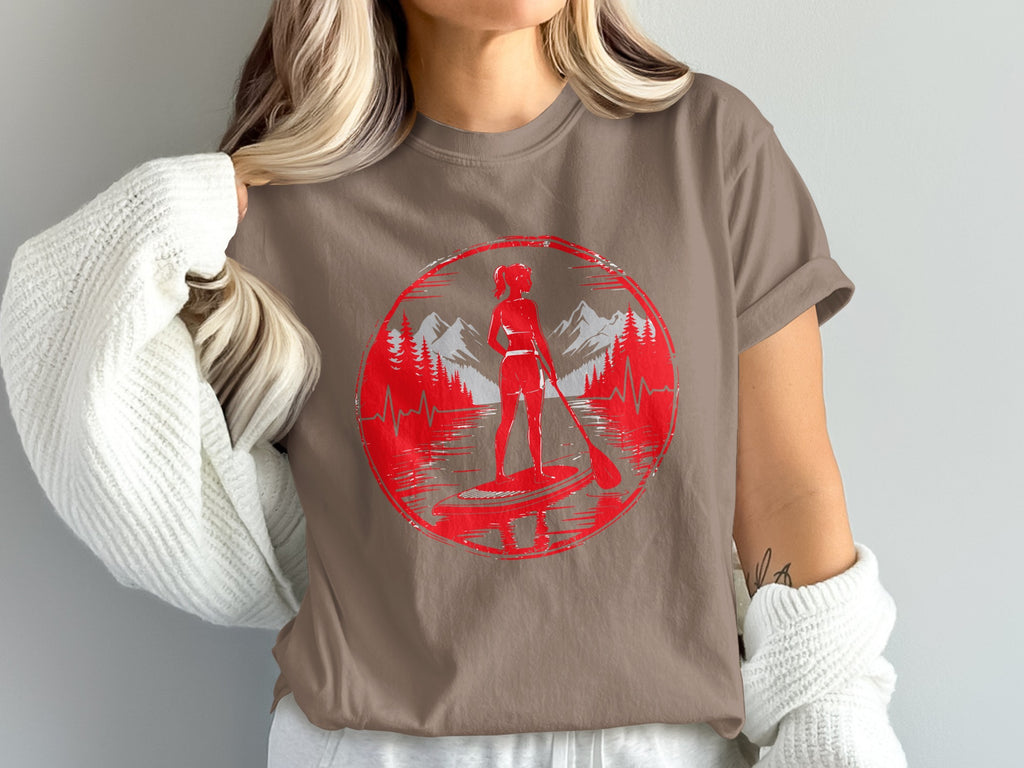 Woman wearing a graphic t-shirt with a stand-up paddleboarder in a mountain landscape.