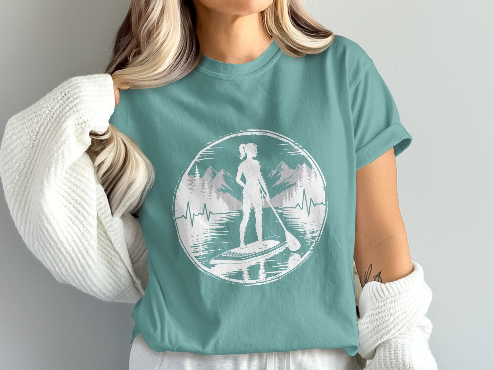 Woman wearing teal t-shirt with paddleboarder graphic and mountain silhouette.
