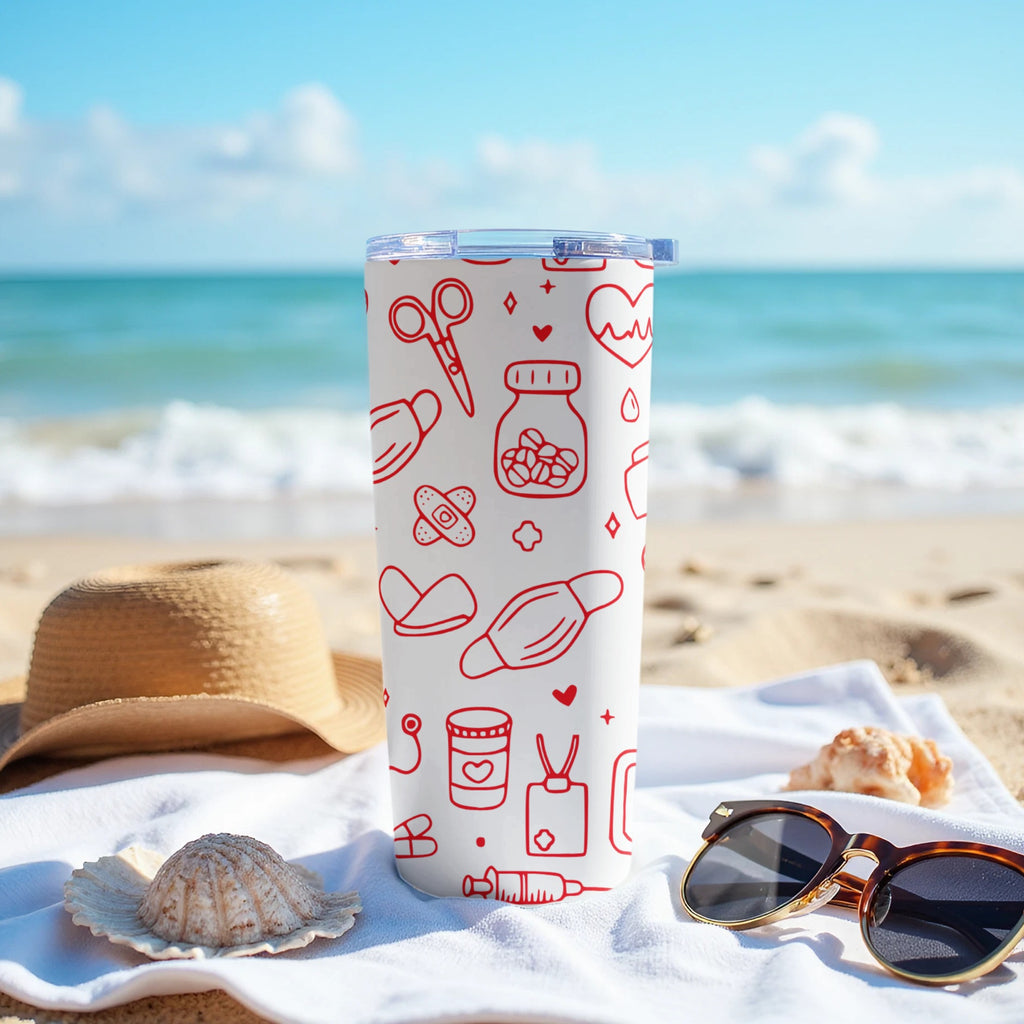 White tumbler with red medical icons on a sandy beach beside sunglasses and a hat.